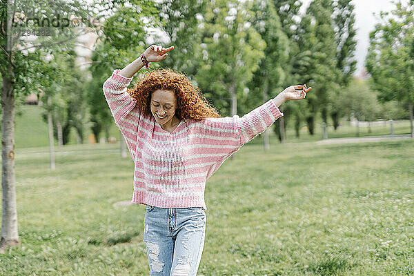 Fröhliche junge rothaarige Frau tanzt mit erhobenen Armen im Park