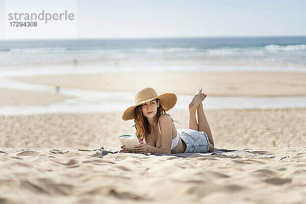 Junge Frau mit Buch  die wegschaut  während sie am Strand im Urlaub auf dem Sand liegt
