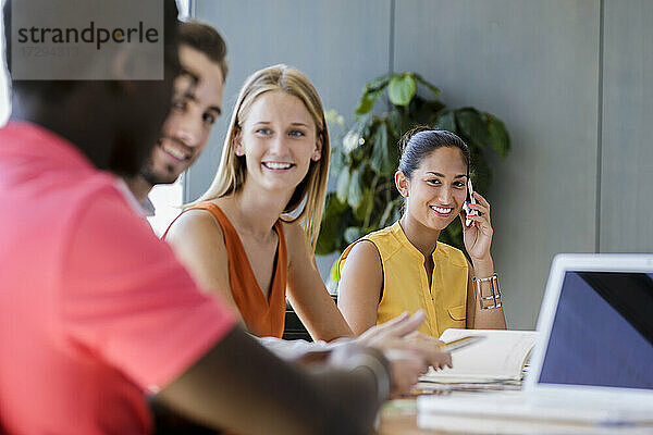 Lächelnde Geschäftsfrau  die auf einem Smartphone spricht  während männliche und weibliche Unternehmer in einer Besprechung im Büro diskutieren