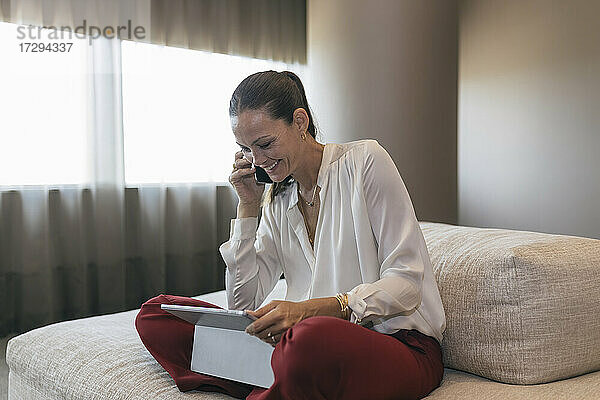 Glückliche reife Geschäftsfrau  die mit einem digitalen Tablet auf dem Sofa sitzt und telefoniert