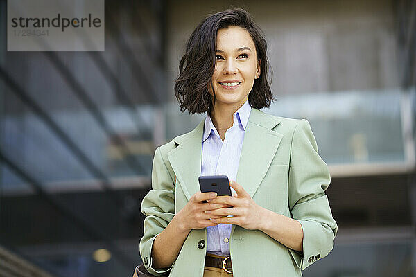 Lächelnde Geschäftsfrau  die wegschaut und ihr Smartphone vor einem Bürogebäude hält