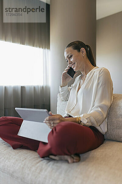 Lächelnde weibliche Fachkraft  die mit einem Smartphone spricht und mit einem digitalen Tablet auf dem Sofa sitzt