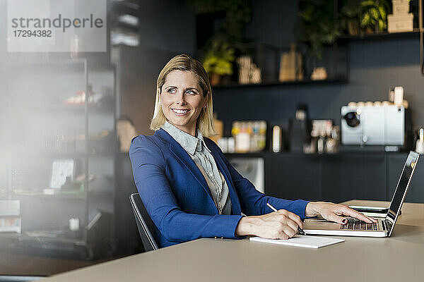 Lächelnde Unternehmerin sitzt mit Laptop am Schreibtisch und schaut weg