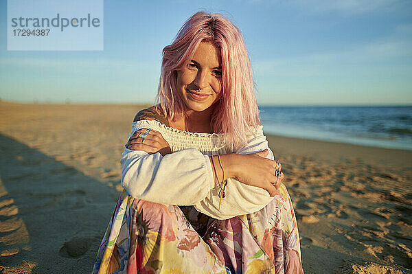 Lächelnde junge Frau mit verschränkten Armen  die bei Sonnenuntergang am Strand sitzt