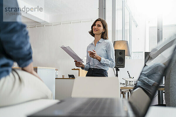 Junge Geschäftsfrau hält Dokumente in der Hand  während sie mit einem Kollegen im Büro diskutiert