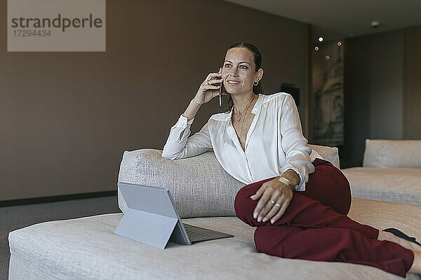 Lächelnde reife Geschäftsfrau  die auf dem Sofa sitzend mit einem Mobiltelefon spricht