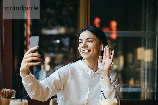 Fröhliche Frau winkt bei einem Videoanruf  während sie in einem Cafe sitzt