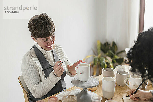 Lächelnde Künstlerin  die eine Tasse bemalt  während sie mit einem Kollegen in einem Workshop arbeitet