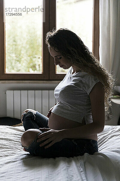 Junge schwangere Frau betrachtet ihren Bauch  während sie auf dem Bett sitzt