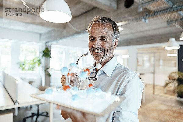 Lächelnder Geschäftsmann hält digitale Tablette und künstliches Molekül im Büro