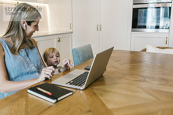 Lächelnde Freiberuflerin mit Blick auf den neugierigen Sohn vor dem Laptop am Esstisch in der Küche