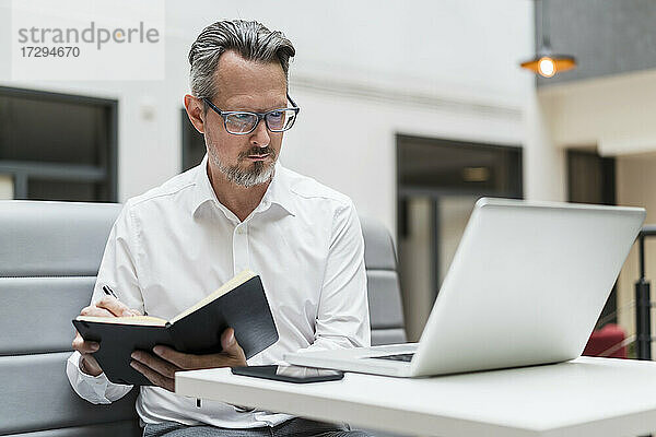 Männlicher Unternehmer mit Buch  der im Büro auf seinen Laptop schaut