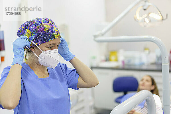 Zahnärztin beim Entfernen der Gesichtsschutzmaske mit Patient im Hintergrund in einer medizinischen Klinik