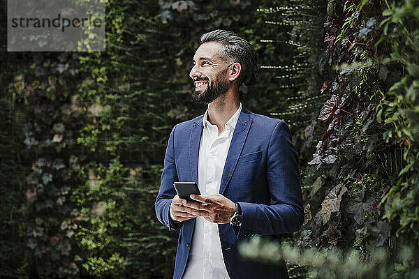 Männlicher Unternehmer mit Mobiltelefon  der lächelnd neben Pflanzen steht