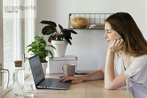 Weibliche Fachkraft mit Hand am Kinn arbeitet am Laptop im Büro zu Hause
