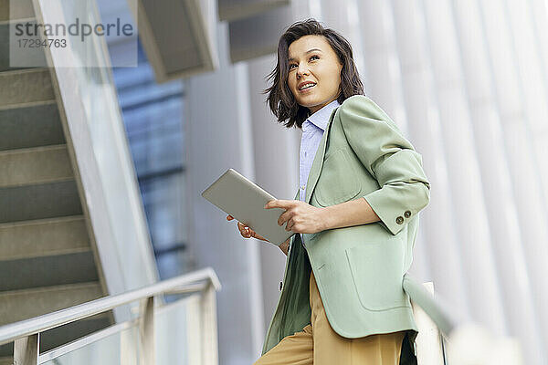Junge Geschäftsfrau mit digitalem Tablet  die den Blick abwendet und sich auf ein Geländer stützt