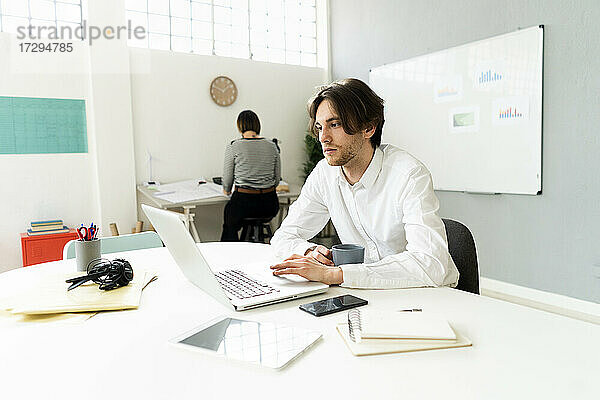 Männlicher Unternehmer  der einen Laptop benutzt  während er mit einem Kollegen im Hintergrund im Büro sitzt