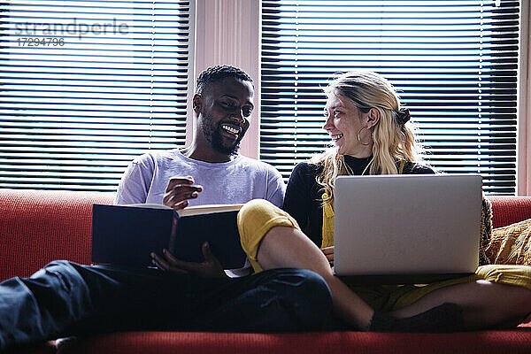 Lächelndes Paar mit Buch und Laptop im Gespräch auf dem Sofa sitzend