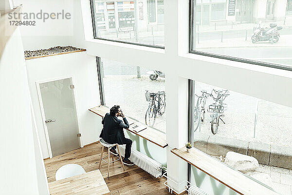 Nachdenklicher männlicher Unternehmer  der mit der Hand am Kinn sitzt und durch ein Fenster im Büro schaut