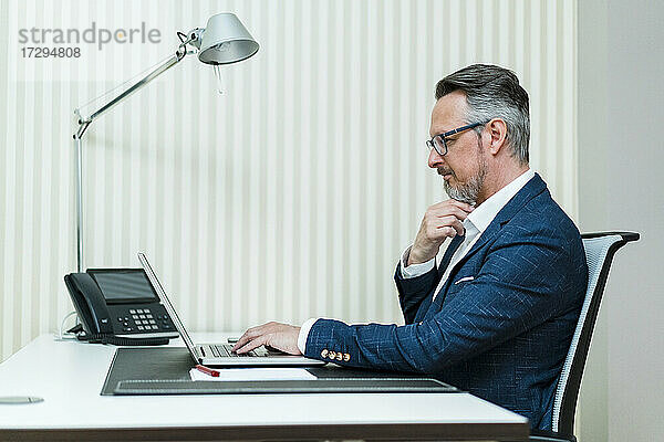 Männlicher Unternehmer mit Hand am Kinn  der einen Laptop am Schreibtisch benutzt