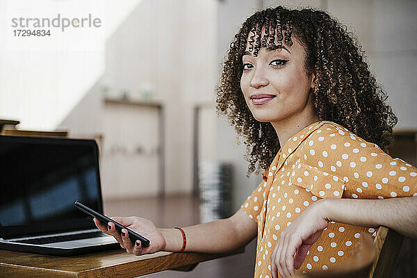 Junge Freiberuflerin hält ihr Smartphone in der Hand  während sie in einem Café am Laptop sitzt