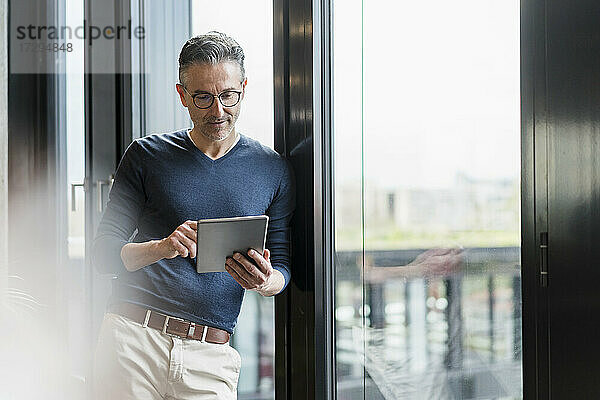 Männlicher Unternehmer  der ein digitales Tablet benutzt und sich an ein Glasfenster im Büro lehnt