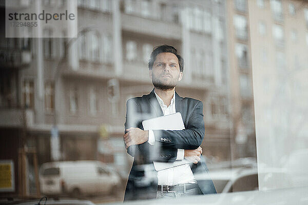 Nachdenklicher männlicher Unternehmer  der ein digitales Tablet hält und durch ein Bürofenster schaut