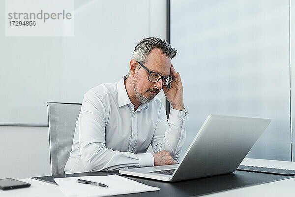 Geschäftsmann mit Kopf in der Hand sitzt mit Laptop am Schreibtisch