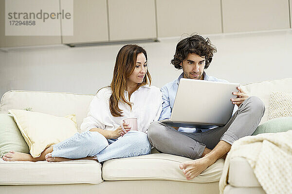 Mid adult man using laptop while girlfriend sitting besides on sofa in new home