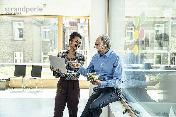 Lächelnde männliche und weibliche Kollegen  die sich gegenseitig ansehen  während sie über einen Laptop im Büro diskutieren