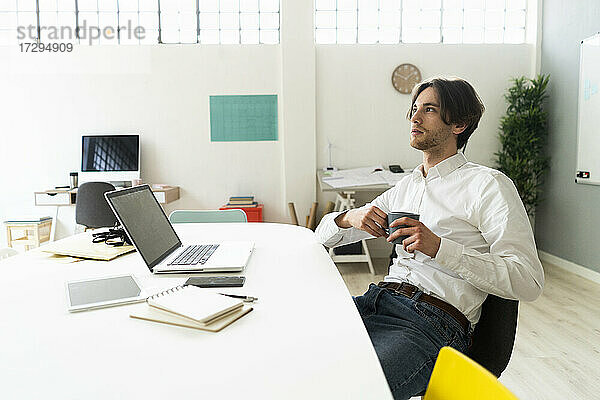 Nachdenklicher Geschäftsmann mit Kaffeetasse am Schreibtisch sitzend im Büro