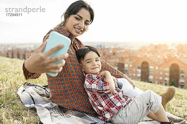Lächelnde Frau  die ein Selfie mit einem Jungen macht  während sie auf einem Hügel sitzt