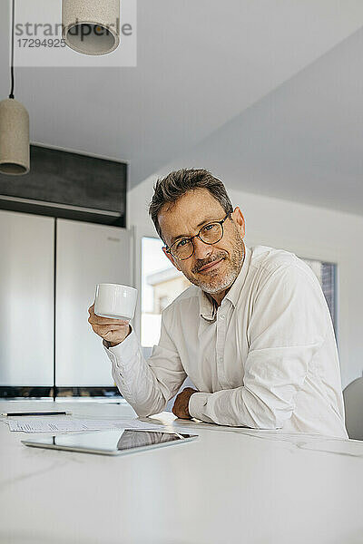 Geschäftsmann mit Brille  der eine Kaffeetasse an der Kücheninsel im Heimbüro hält