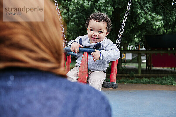 Lächelnder Junge schaut Frau an  während er auf einer Schaukel auf dem Spielplatz sitzt