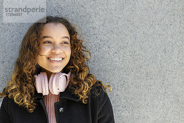 Lächelnde junge Frau mit Kopfhörern  die vor einer Wand nachdenkt