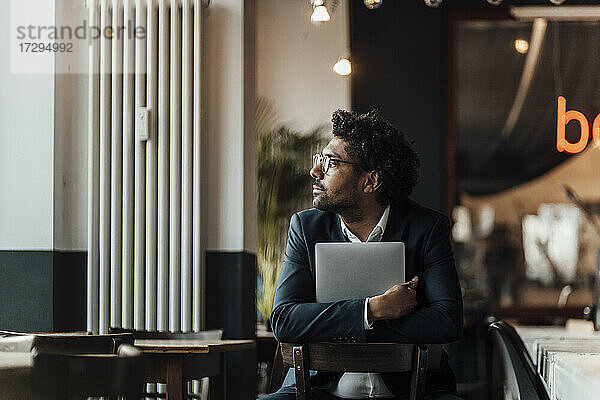 Männlicher Unternehmer  der einen Laptop hält und auf einem Stuhl in einem Café sitzt und wegschaut