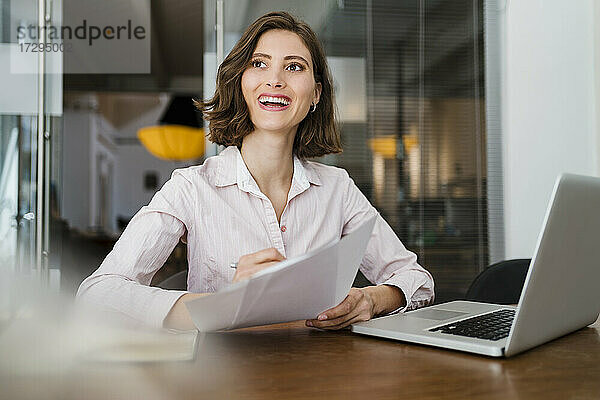 Schöne lächelnde Unternehmerin sitzt mit Dokument am Schreibtisch und schaut weg