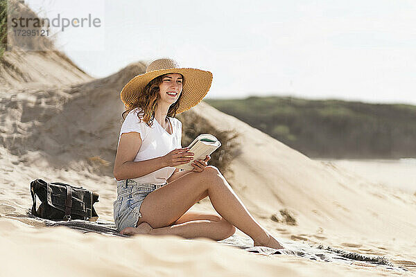 Lächelnde junge Frau mit Hut sitzt mit Buch an einem sonnigen Tag am Strand während der Ferien