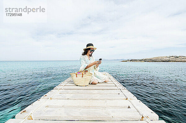 Lächelnde Frau  die ein Mobiltelefon benutzt  während sie im Urlaub auf einem Steg sitzt