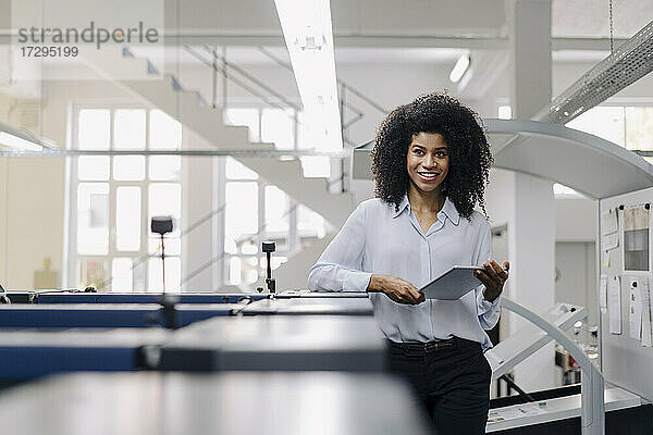 Lächelnde Afro-Geschäftsfrau mit digitalem Tablet  die sich auf eine Maschine in der Industrie stützt