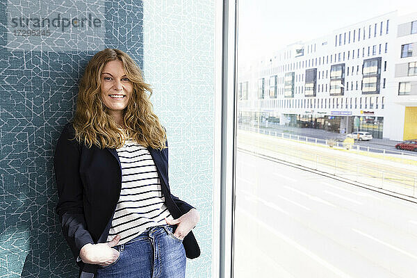 Weibliche Unternehmerin lächelt  während sie sich im Büro an die Wand am Fenster lehnt