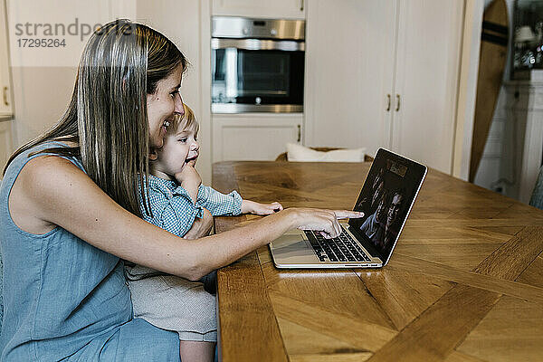 Lächelnde Frau  die auf einen Laptop zeigt  während ihr Sohn während eines Videogesprächs mit der Familie in der Küche auf dem Schoß sitzt