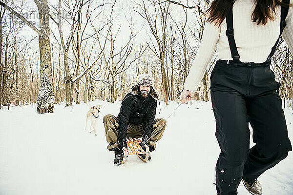 Frau zieht Mann auf Holzschlitten sitzend mit Hund im Wald hinter sich her