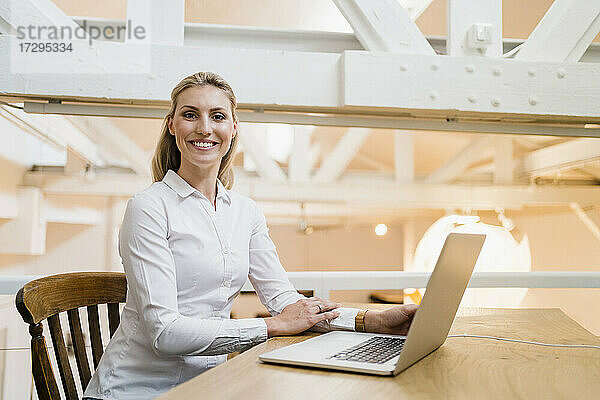 Lächelnde blonde Unternehmerin sitzt mit Laptop in einem kreativen Büro