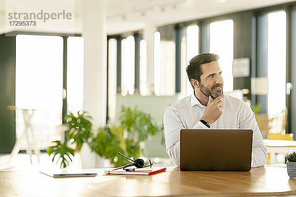 Geschäftsmann mit Hand am Kinn  der vor einem Laptop im Büro sitzt und nachdenkt