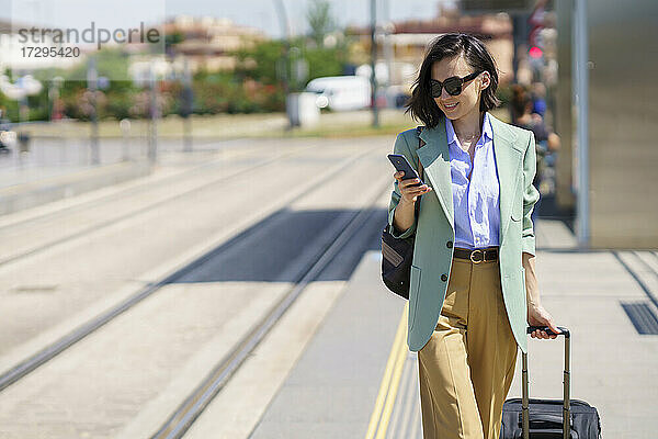 Berufstätige Frau  die ein Smartphone benutzt  während sie mit einem Koffer am Bahnhof spazieren geht