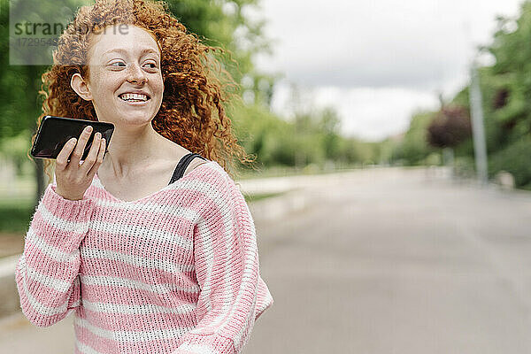 Schöne lächelnde rothaarige Frau  die im Park über einen Lautsprecher mit ihrem Handy spricht und dabei wegschaut