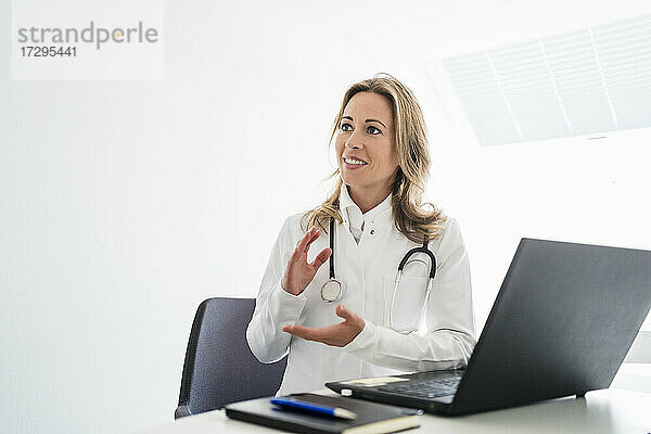 Ärztin schaut weg und gestikuliert während einer Videokonsultation am Schreibtisch