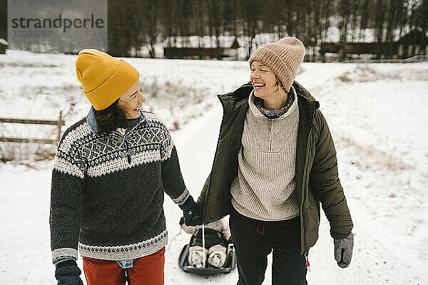 Lächelndes lesbisches Paar schaut sich gegenseitig an  während es seine Tochter durch den Schnee zieht