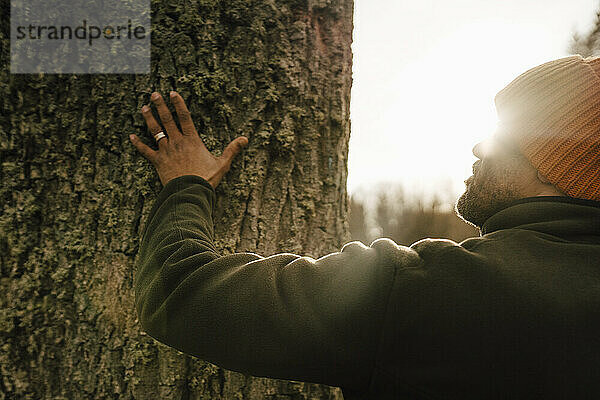 Älterer männlicher Wanderer berührt Baumstamm im Wald an einem sonnigen Tag